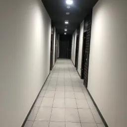 Korytarz biurowy z białymi ścianami, szarą podłogą z płytek i czarnym sufitem z rzędem okrągłych lamp, widoczne drzwi z okleiną drewnopodobną, zabezpieczone folią.