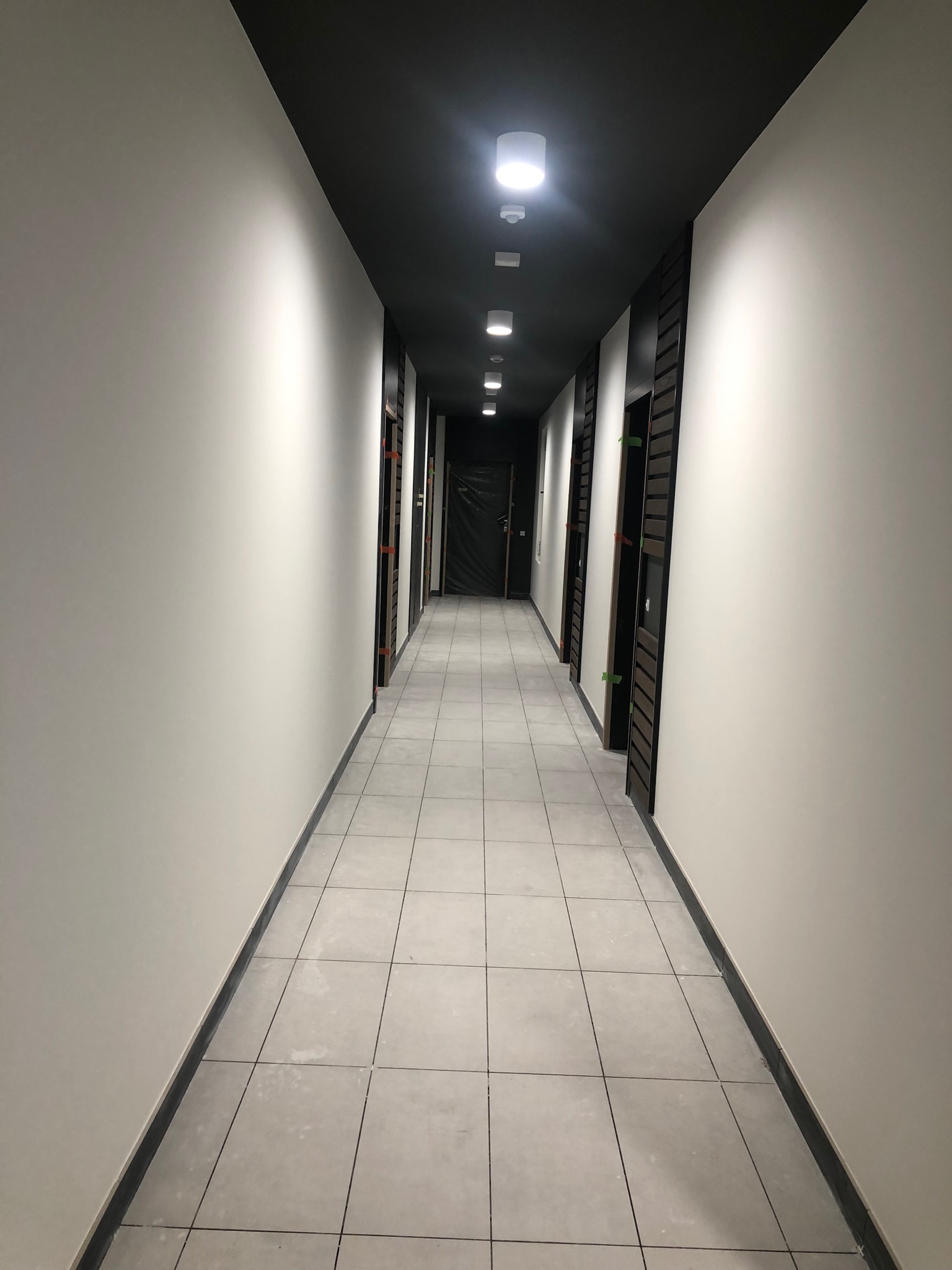 Korytarz biurowy z białymi ścianami, szarą podłogą z płytek i czarnym sufitem z rzędem okrągłych lamp, widoczne drzwi z okleiną drewnopodobną, zabezpieczone folią.