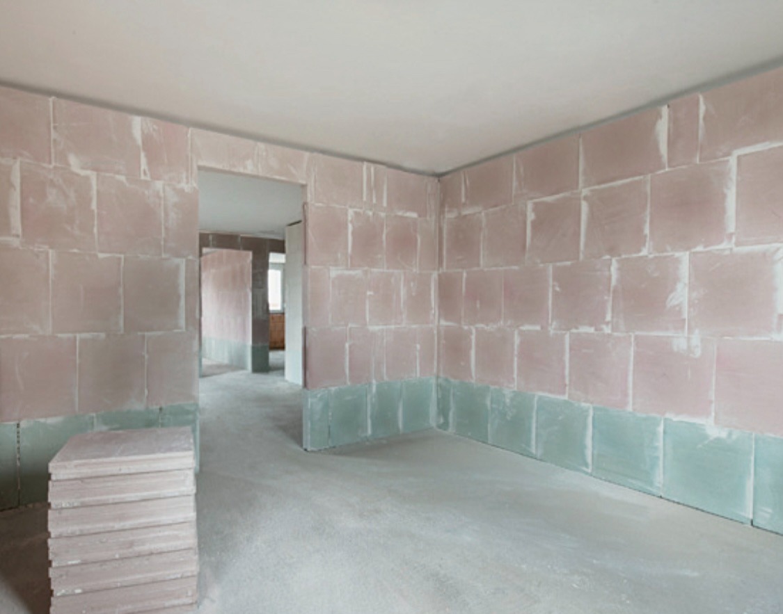 Wnętrze w trakcie prac murarskich: ściany z bloków, częściowo pokryte zaprawą, widoczne przejście do innego pomieszczenia, stos bloków na podłodze.