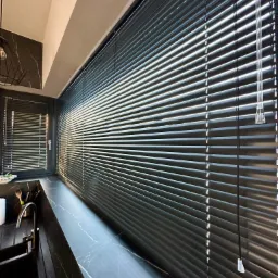 Ciemne żaluzje aluminiowe w oknie kuchennym, z widocznym fragmentem blatu i zlewu z czarnego marmuru.