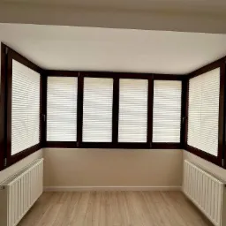 Wnętrze pokoju z trzema oknami wyposażonymi w jasne, poziome osłony okienne w ciemnych, drewnianych ramach, grzejniki pod oknami, panele podłogowe.