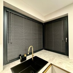 Roll Design - Nowoczesna kuchnia z czarnym zlewem i złotą armaturą, okna z szarymi żaluzjami aluminiowymi w czarnych ramach, marmurowy blat.