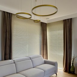 Roll Design - Jasne żaluzje poziome w nowoczesnym salonie z beżową sofą i złotymi lampami pierścieniowymi, dopełnione brązowymi zasłonami.