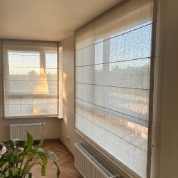 Roll Design - Beżowe rolety rzymskie z delikatnej tkaniny, zamontowane w oknach. Widoczne promienie słoneczne wpadające do wnętrza, podkreślające fakturę materiału.