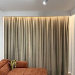 Roll Design - Beżowe, gładkie zasłony zakrywające całą ścianę, z delikatnym podświetleniem od góry, widoczne z fragmentem brązowej sofy i poduszki w salonie.