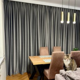 Roll Design - Eleganckie, grafitowe zasłony w salonie z drewnianym stołem i lampami wiszącymi. Na kanapie siedzi pręgowany kot z białym futrem na piersi.