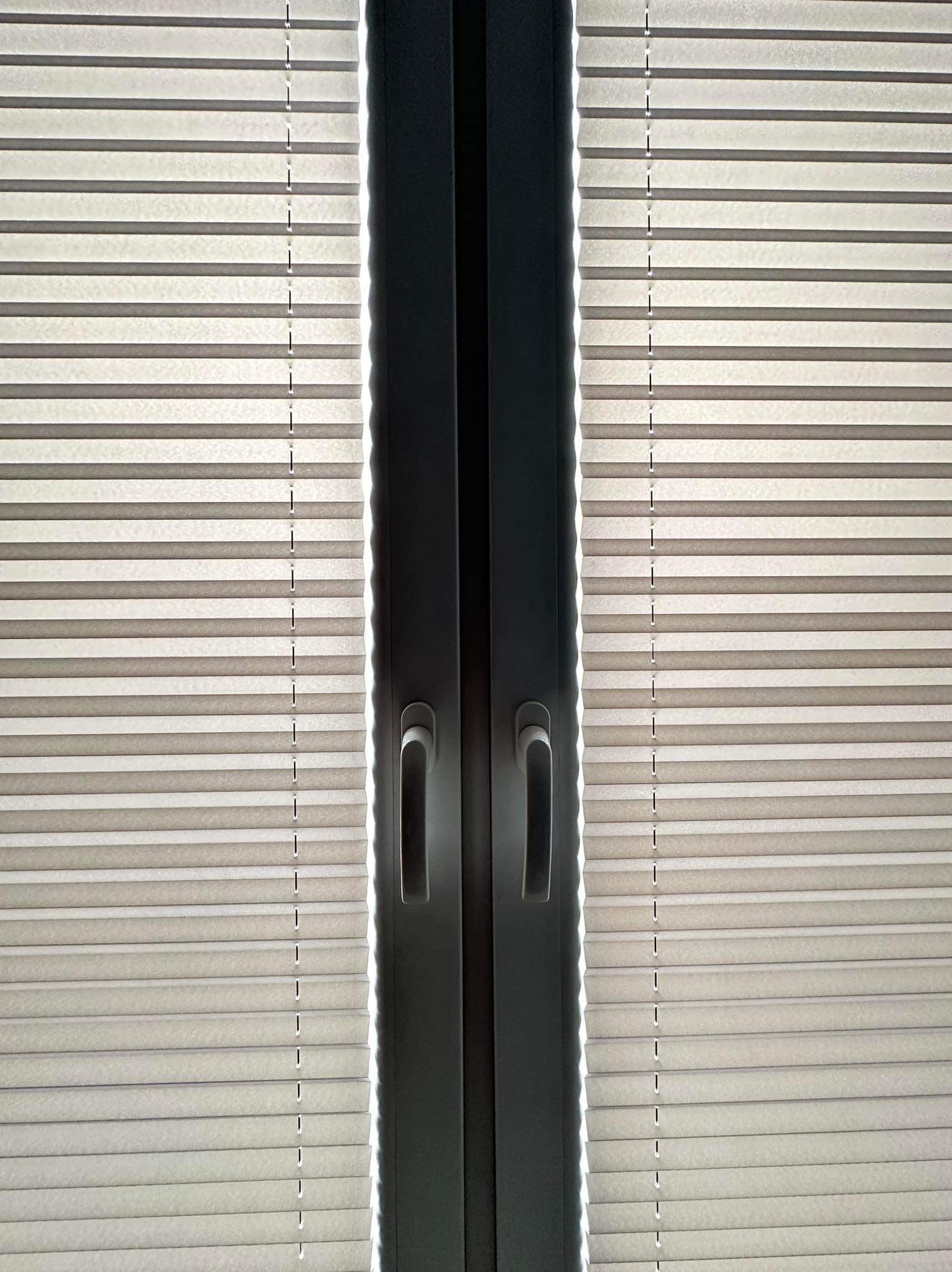 Zamknięte, beżowe plisy okienne z czarnymi uchwytami na oknie balkonowym.