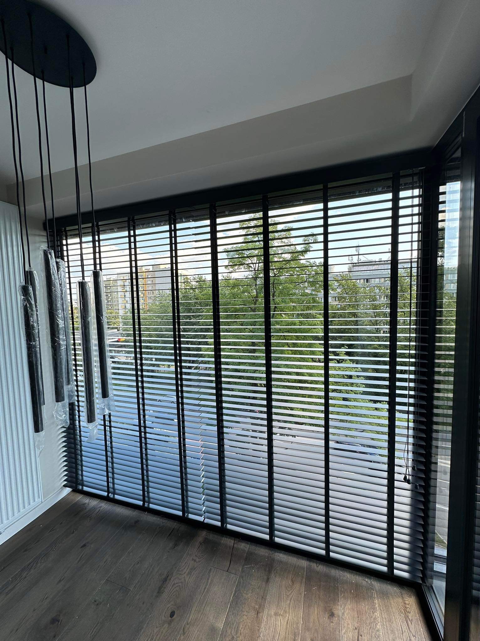 Nowoczesne wnętrze z ciemnymi żaluzjami aluminiowymi i drewnianą podłogą, widok na zieleń za oknem, lampy wiszące w folii ochronnej.