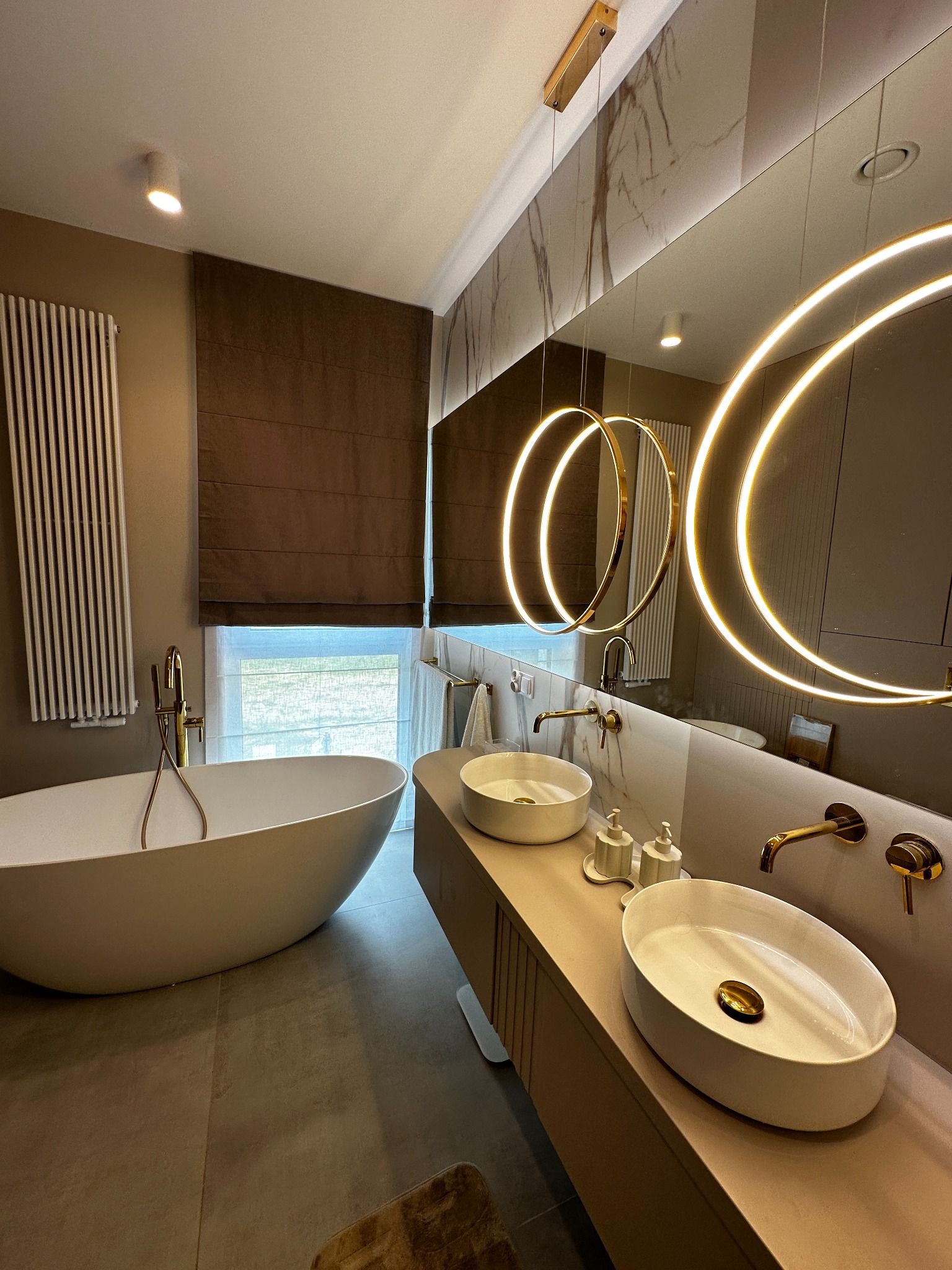 Nowoczesna łazienka z wolnostojącą wanną, brązowymi zasłonami rzymskimi, dwiema umywalkami nablatowymi z bateriami w kolorze złotym i dużym lustrem z okrągłymi lampami LED.