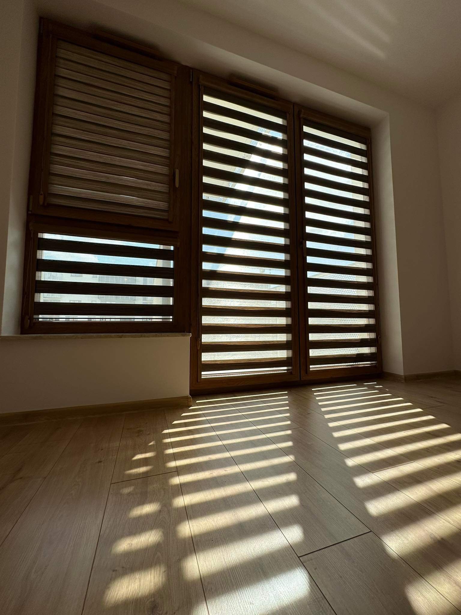 Trzy okna z drewnianymi ramami wyposażone w rolety wewnętrzne typu dzień-noc, rzucające cienie na podłogę z paneli.