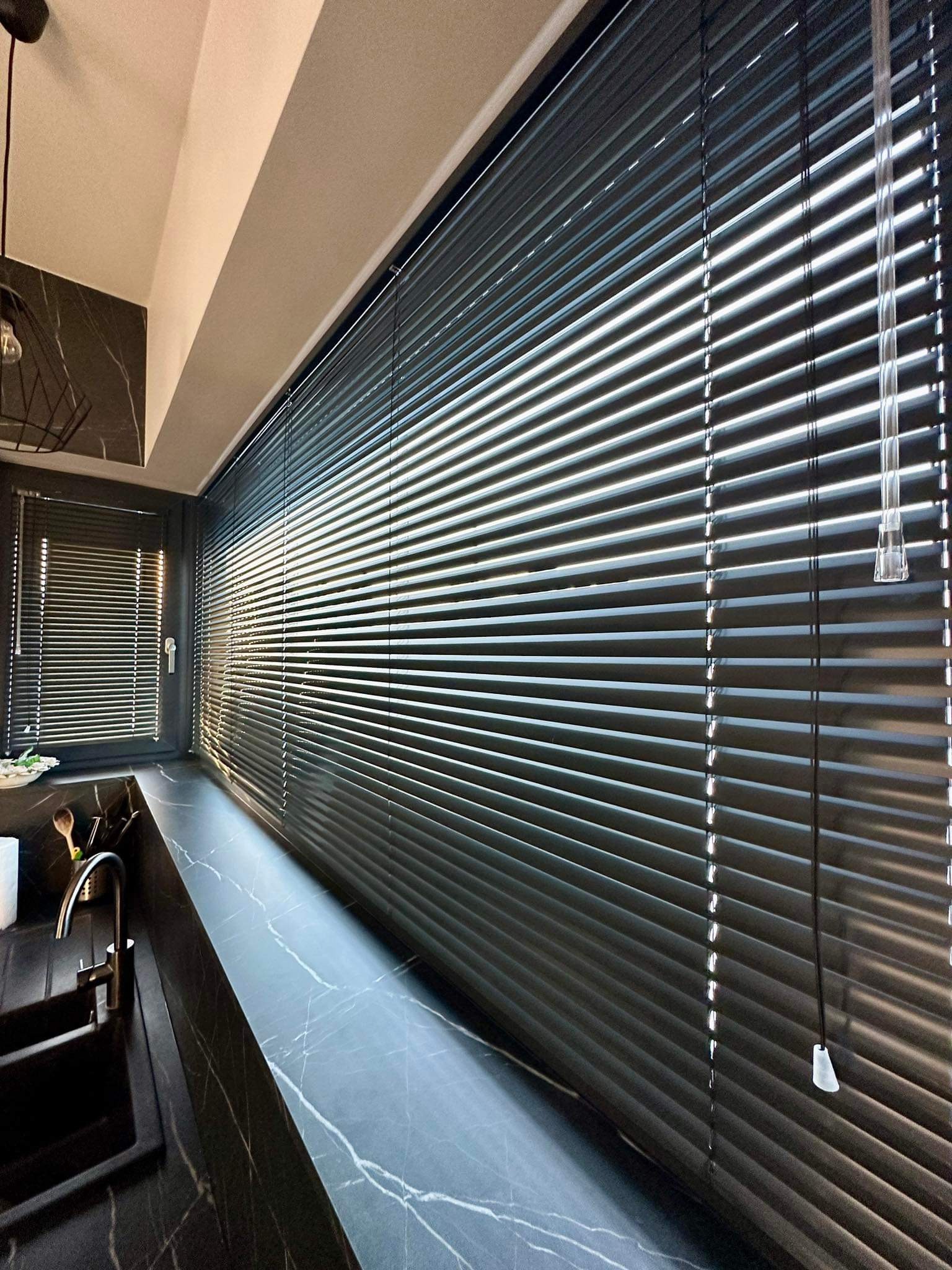 Ciemne żaluzje aluminiowe w oknie kuchennym, z widocznym fragmentem blatu i zlewu z czarnego marmuru.