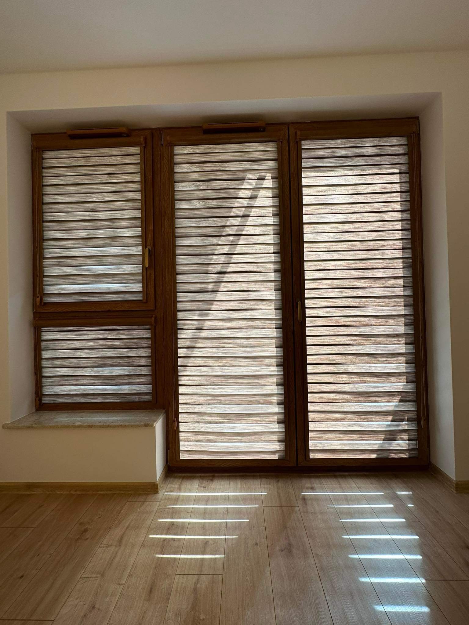 Trzy okna z drewnianymi ramami wyposażone w poziome, dwukolorowe osłony przeciwsłoneczne, przez które wpadają promienie słoneczne na parkiet.