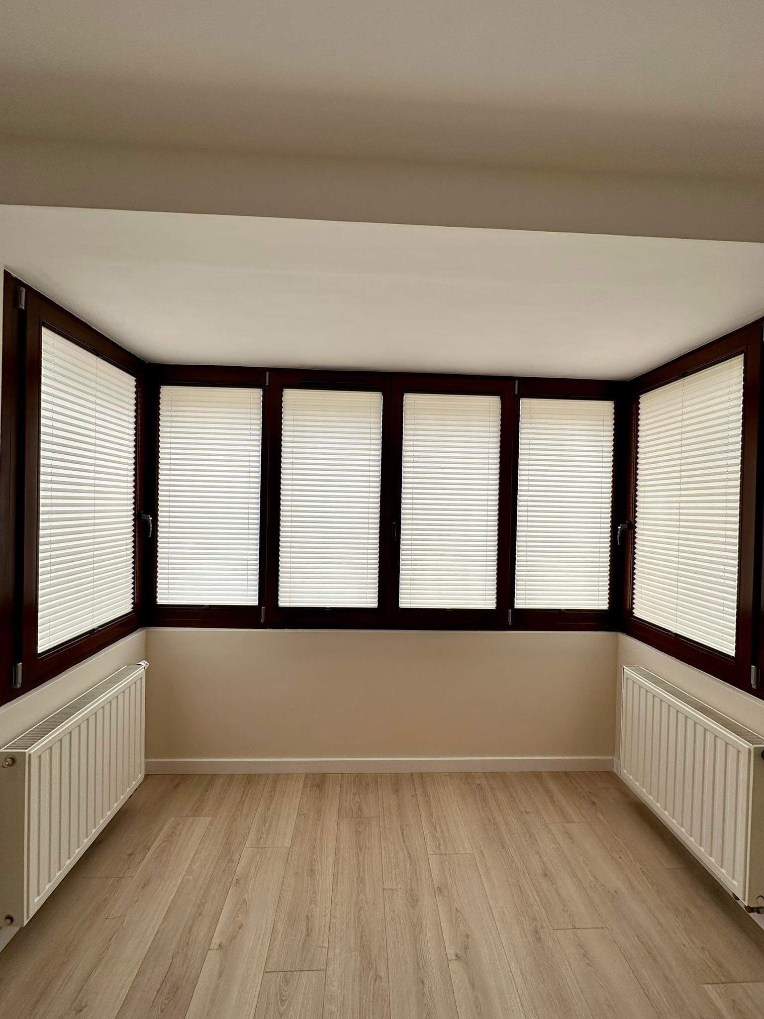 Wnętrze pokoju z trzema oknami wyposażonymi w jasne, poziome osłony okienne w ciemnych, drewnianych ramach, grzejniki pod oknami, panele podłogowe.