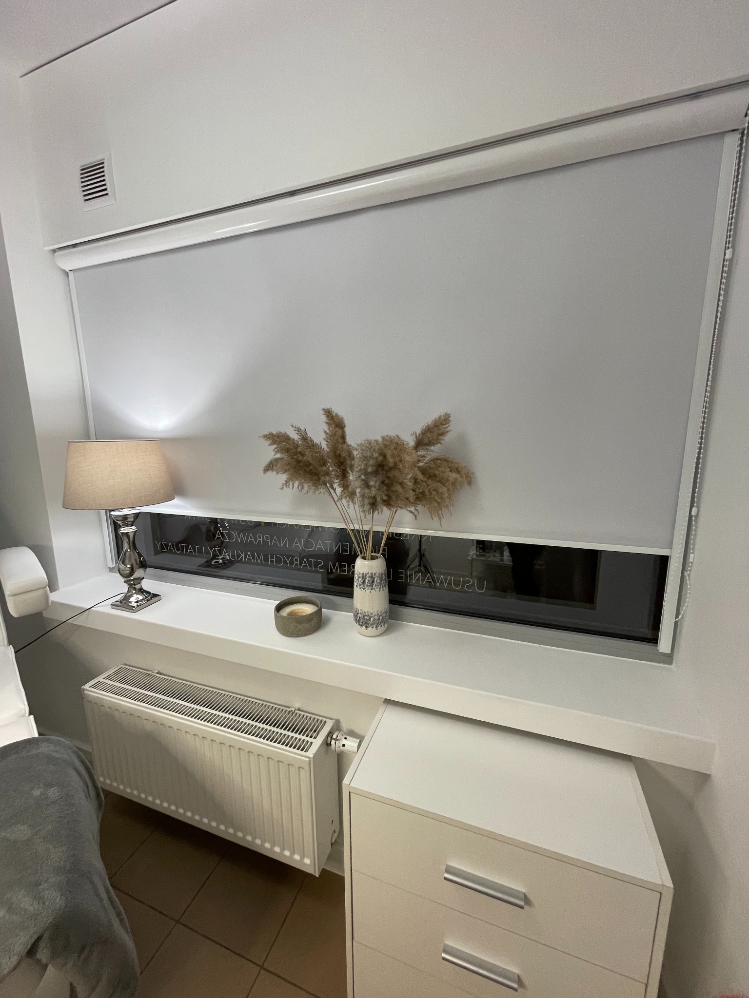 Szara roleta wewnętrzna zwinięta do połowy okna, na parapecie lampka nocna z beżowym abażurem, wazon z suchymi trawami i świeczka, widoczny grzejnik i fragment komody.