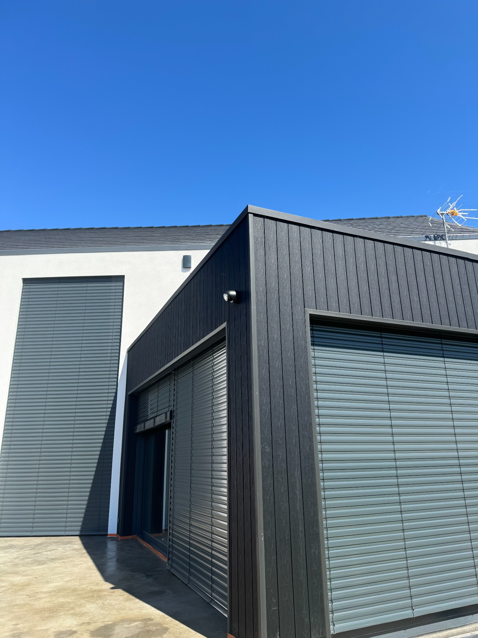 Nowoczesny budynek z szarą elewacją i zewnętrznymi osłonami okiennymi w kolorze szarym, częściowo zasłoniętymi, pod błękitnym niebem, widoczna antena na dachu.