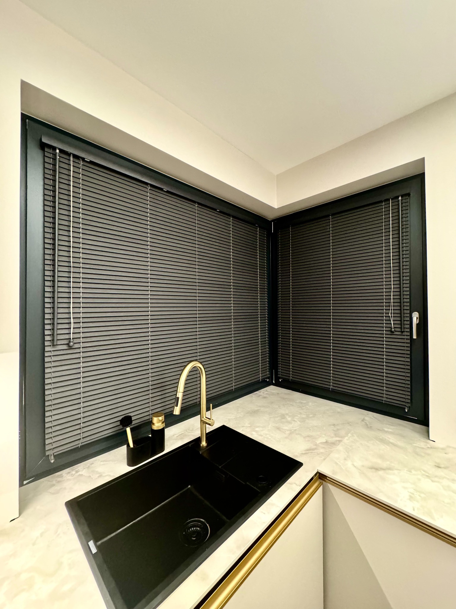 Nowoczesna kuchnia z czarnym zlewem i złotą armaturą, okna z szarymi żaluzjami aluminiowymi w czarnych ramach, marmurowy blat.