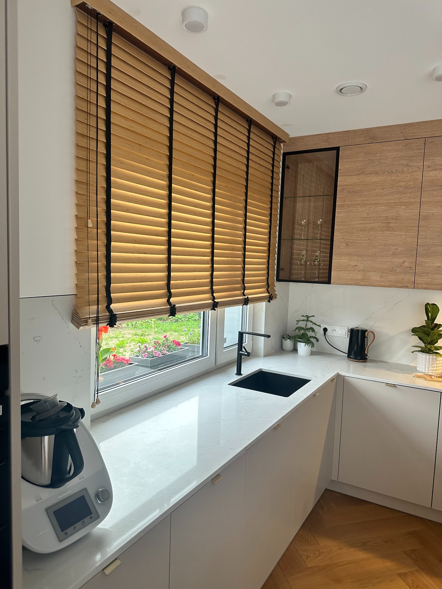 Drewniane żaluzje poziome w kuchni w Jabłonnie, z widokiem na kwiaty w oknie. Naturalne światło podkreśla strukturę drewna i dodaje ciepła wnętrzu.