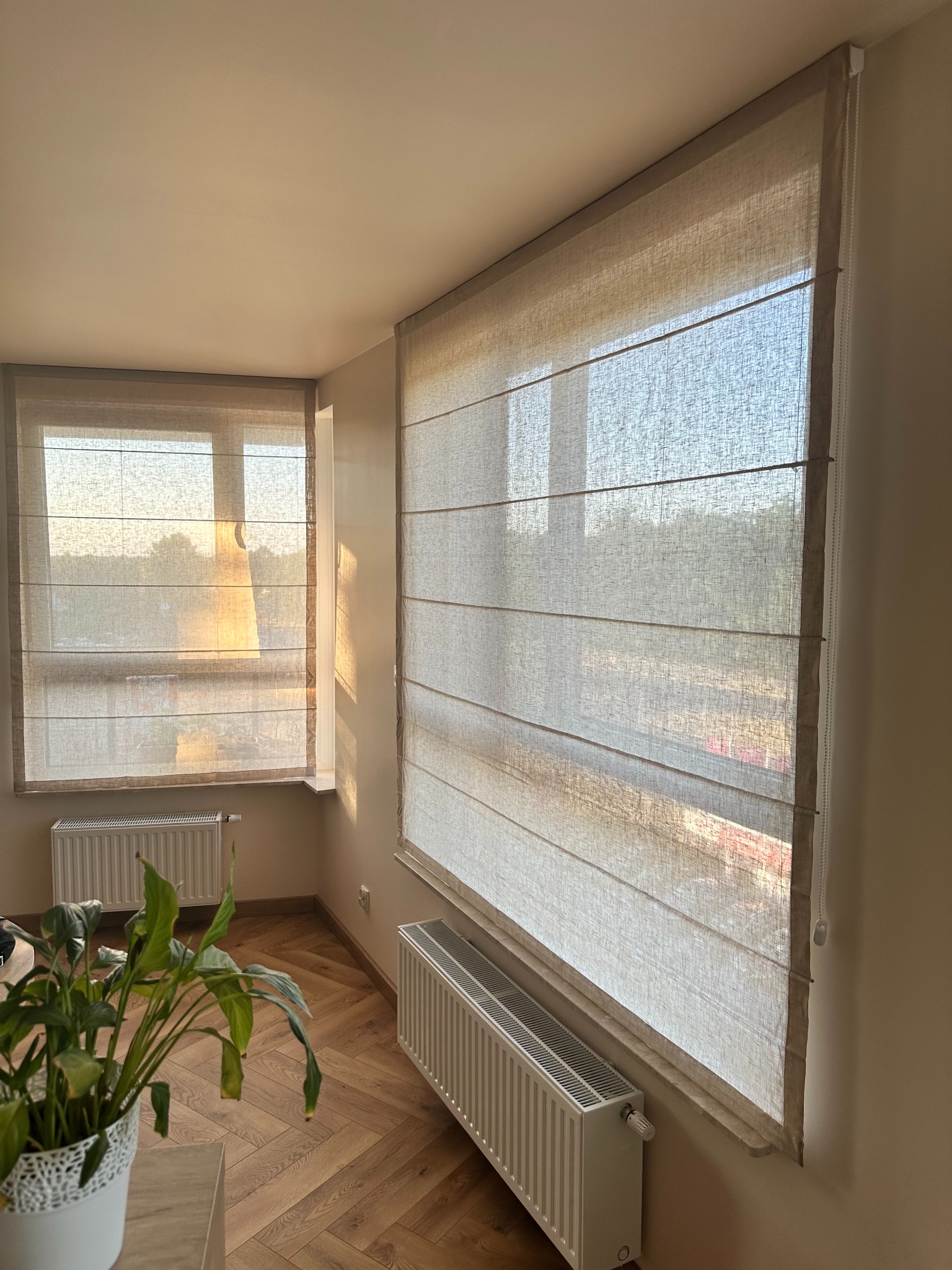 Beżowe rolety rzymskie z delikatnej tkaniny, zamontowane w oknach. Widoczne promienie słoneczne wpadające do wnętrza, podkreślające fakturę materiału.