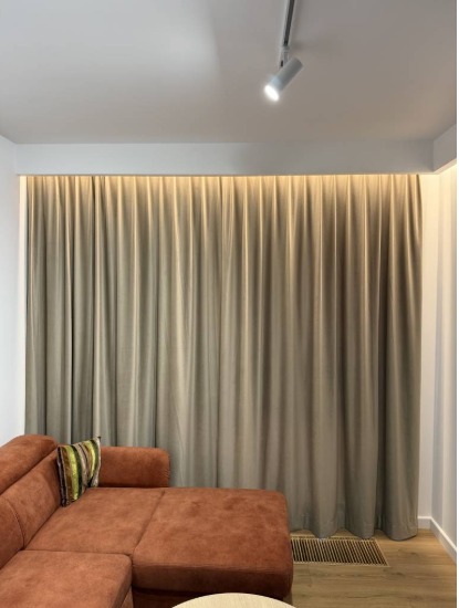 Beżowe, gładkie zasłony zakrywające całą ścianę, z delikatnym podświetleniem od góry, widoczne z fragmentem brązowej sofy i poduszki w salonie.
