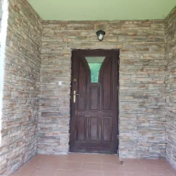 Wejście do domu z elewacją z kamienia dekoracyjnego i brązowymi drzwiami z szybą, oświetlone lampą sufitową.