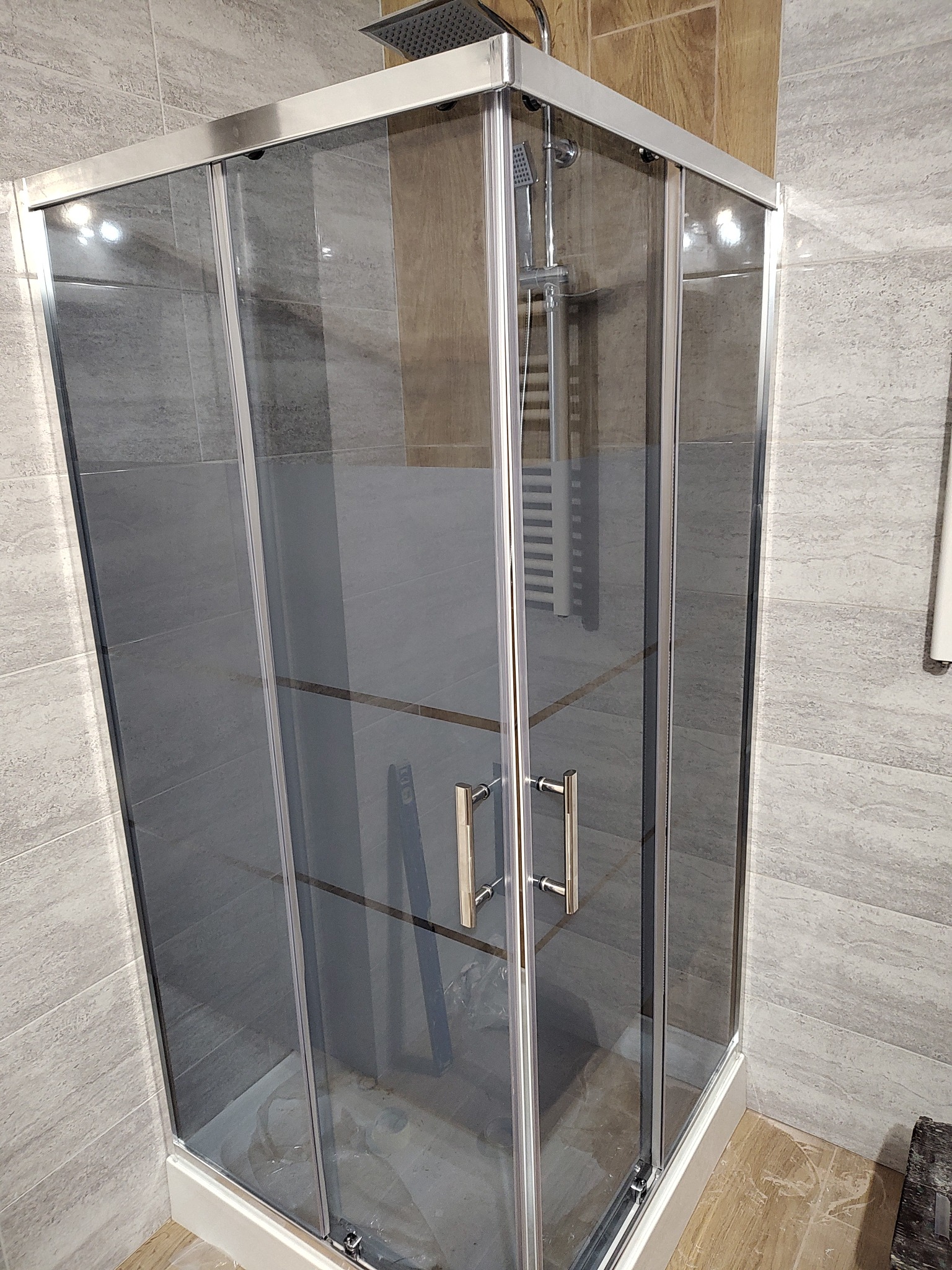 Nowoczesna kabina prysznicowa z przesuwnymi drzwiami i deszczownicą, otoczona płytkami w odcieniach szarości i beżu, częściowo zakryta folią ochronną na podłodze.