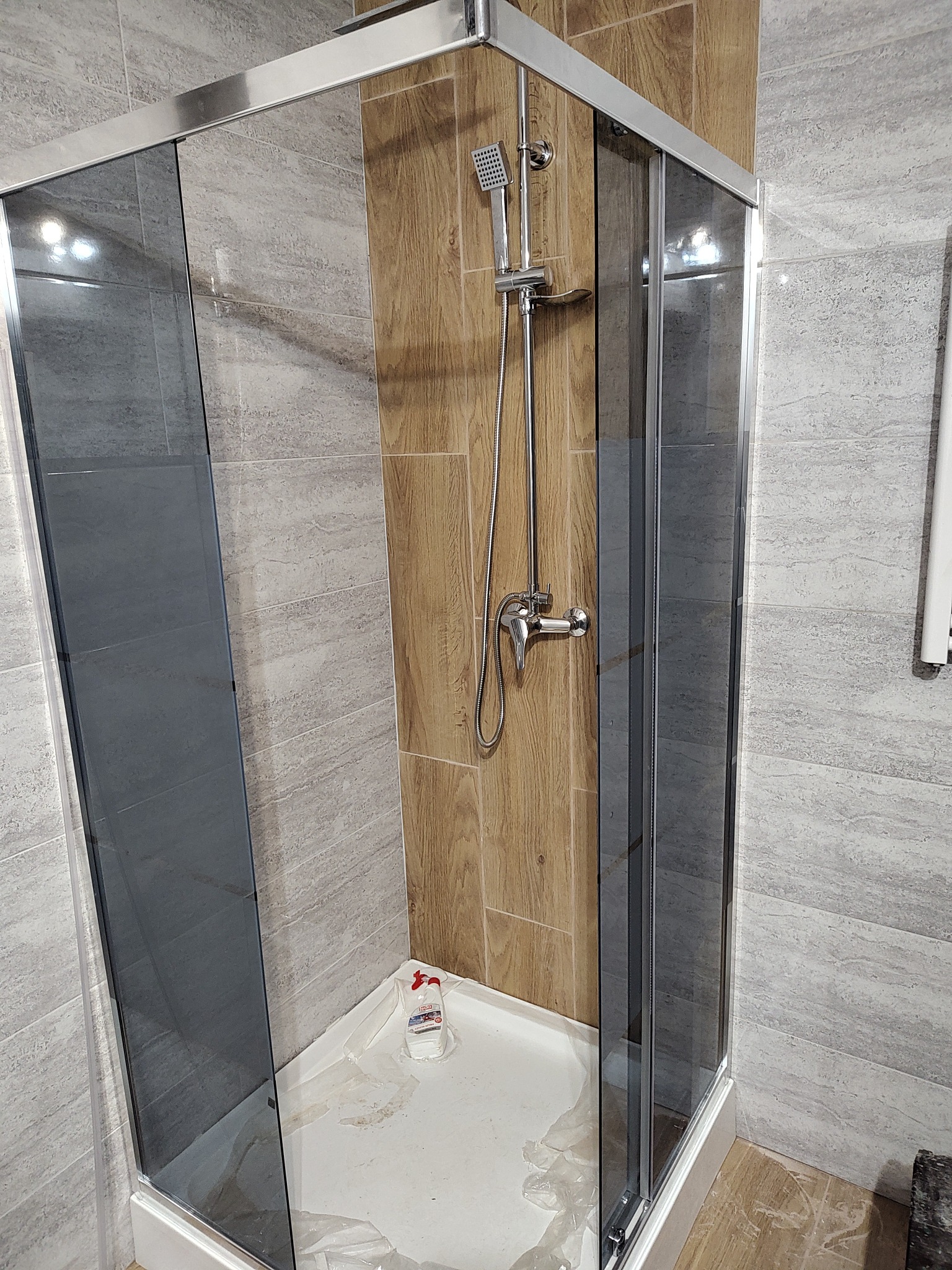 Nowoczesna kabina prysznicowa z szarymi szybami i chromowanymi elementami, zamontowana w łazience wykończonej płytkami imitującymi drewno i szary marmur, z widocznymi pozostałościami po remoncie...