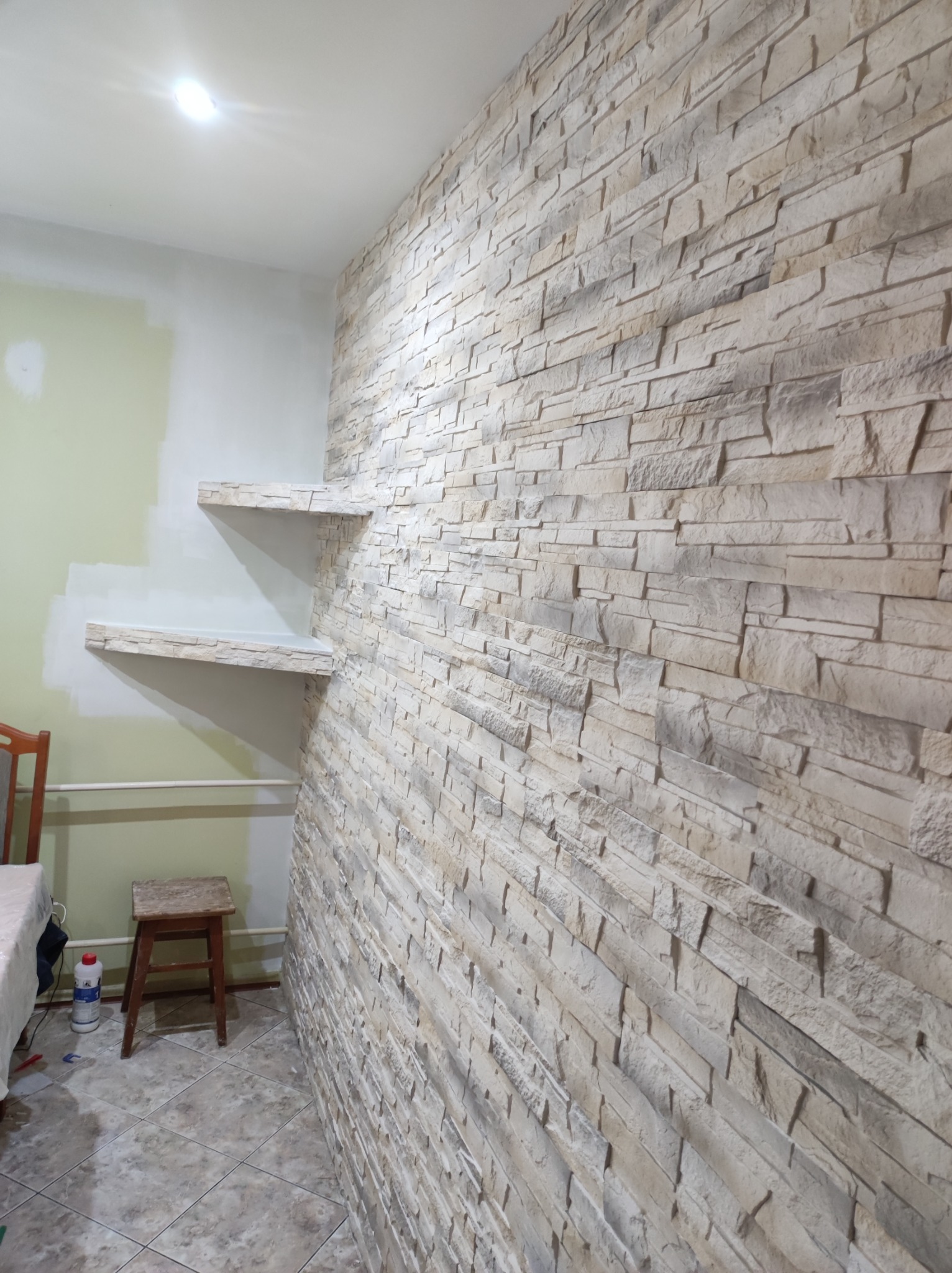 Ściana wyłożona dekoracyjnym kamieniem z piaskowca, z dwiema półkami z tego samego materiału, na tle pomalowanej na zielono ściany, widoczny fragment stołu i stołka.