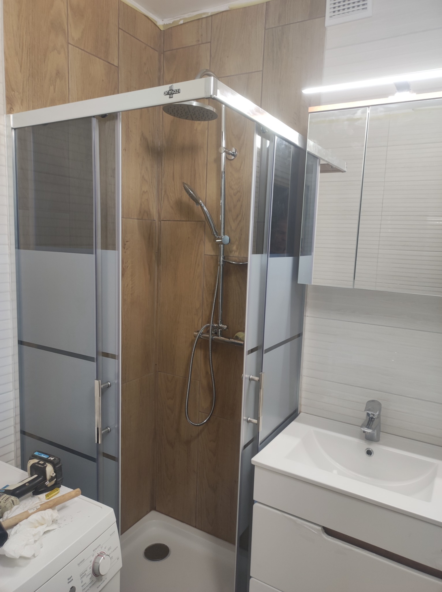 Nowa kabina prysznicowa z deszczownicą, zamontowana w łazience z płytkami imitującymi drewno, widoczna pralka z narzędziami na górze.