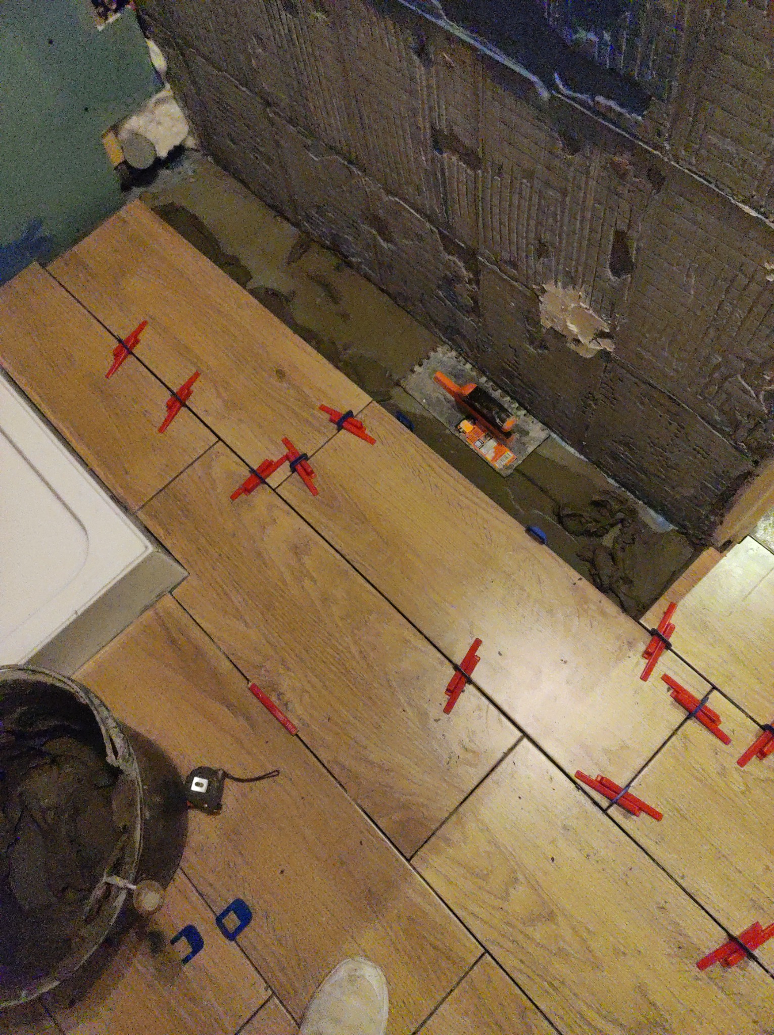 Układanie drewnopodobnych płytek podłogowych w łazience, z użyciem krzyżyków dystansowych i narzędzi glazurniczych, widoczna ściana przygotowana pod płytki.