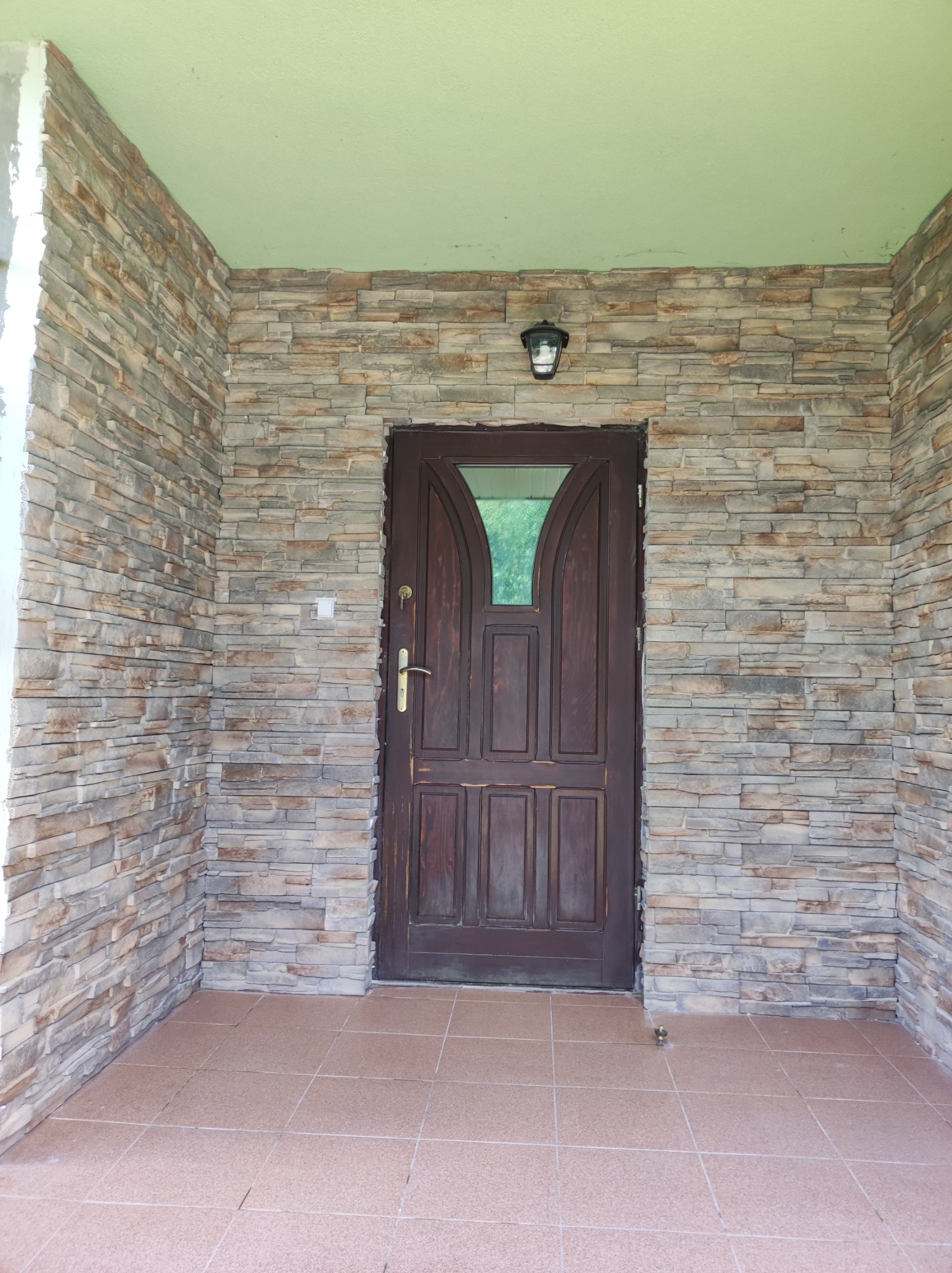 Wejście do domu z elewacją z kamienia dekoracyjnego i brązowymi drzwiami z szybą, oświetlone lampą sufitową.