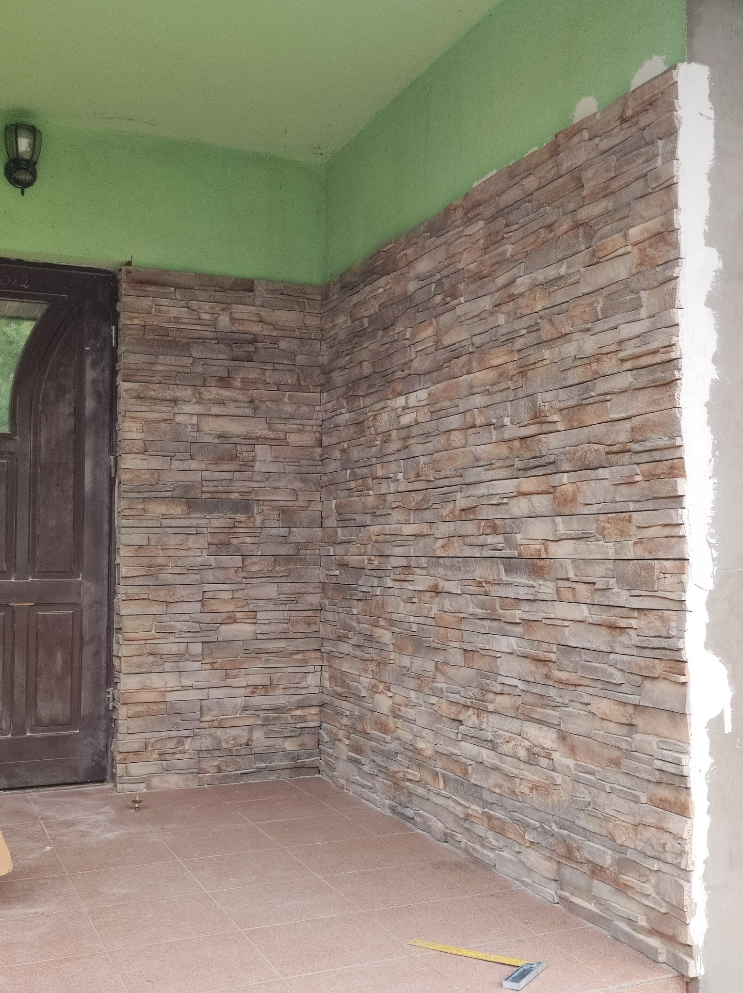 Montaż okładziny kamiennej na ścianie przy wejściu, widoczny fragment elewacji z brązową stolarką drzwiową, zielony sufit i płytki podłogowe, w tle fragment ściany w trakcie wykończenia.