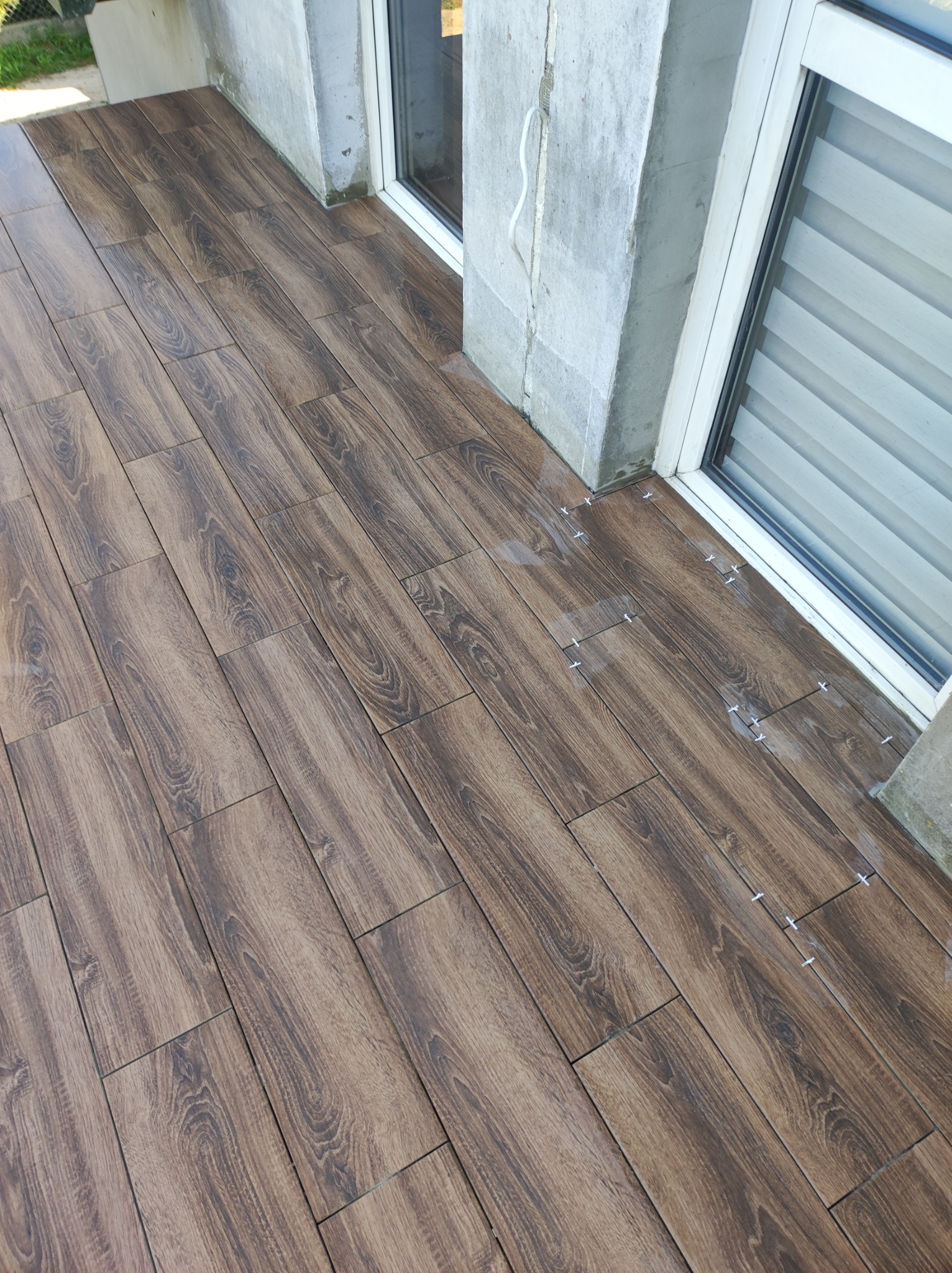 Ułożone płytki imitujące drewno na balkonie, z widocznymi krzyżykami dystansowymi przy oknach.