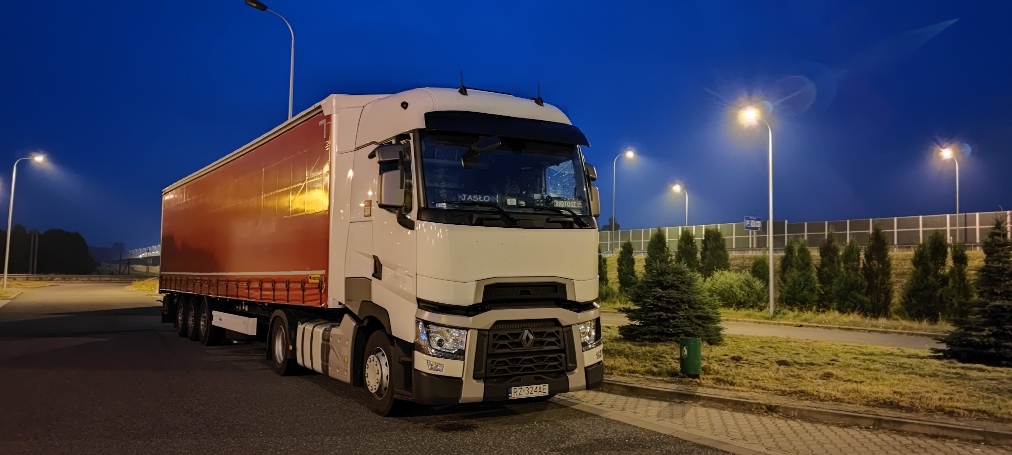 Ciężarówka Renault z naczepą na parkingu o zmierzchu, oświetlona lampami ulicznymi. Na przedniej szybie widoczne napisy 'JAŚLO' i 'BARTOSZ'.