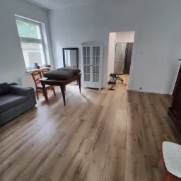 Jasne panele podłogowe w salonie w Gdańsku, widok na szarą sofę, stół z krzesłami i szafę. W tle widoczny odkurzacz i lodówka w kuchni.