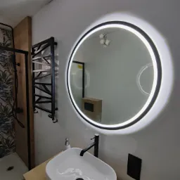 Nowoczesna łazienka z okrągłym podświetlanym lustrem, białą umywalką nablatową z czarną armaturą, czarnym grzejnikiem drabinkowym i kabiną prysznicową z motywem liści.