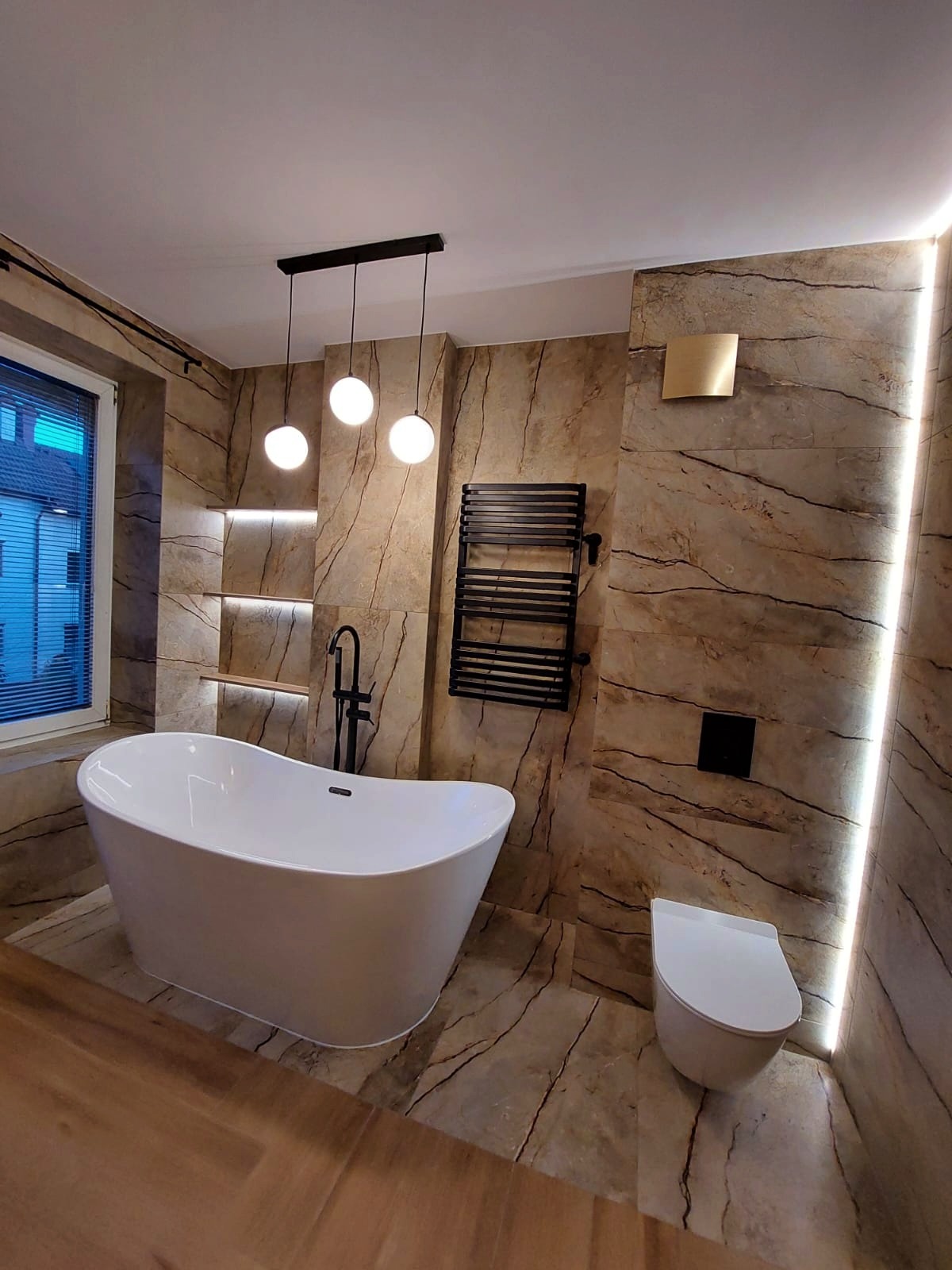 Nowoczesna łazienka z wolnostojącą białą wanną, marmurowymi płytkami, czarną armaturą, grzejnikiem drabinkowym i oświetleniem LED wzdłuż ściany.