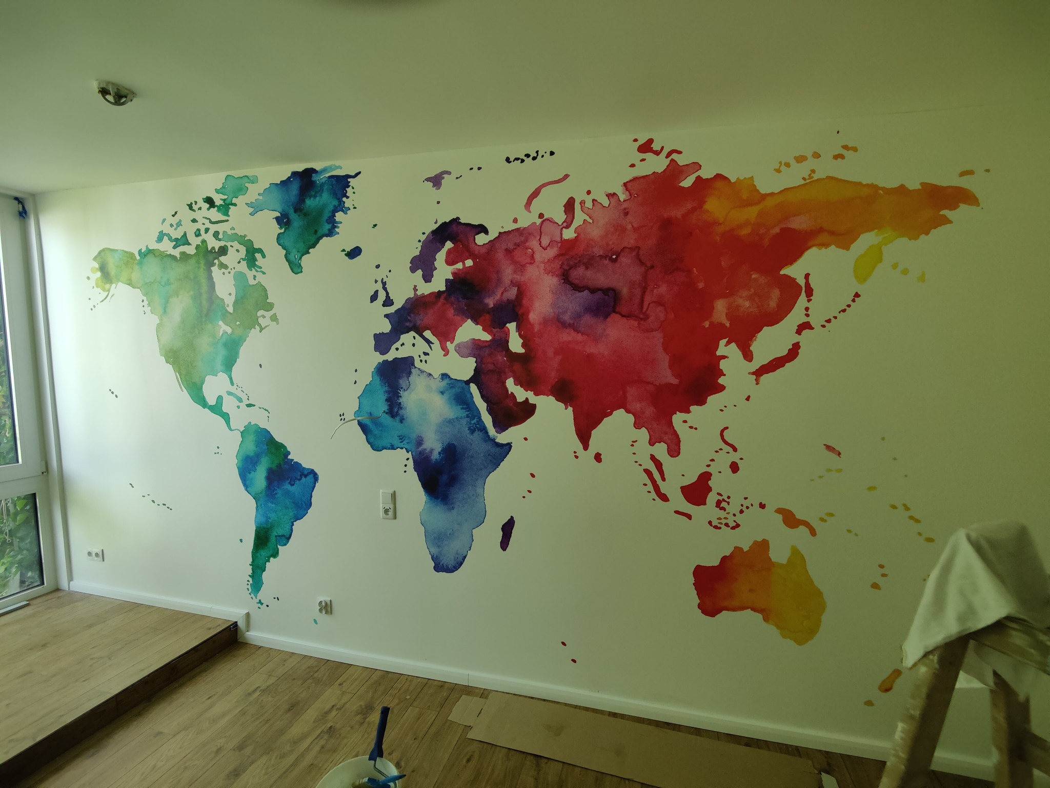 Ściana z ręcznie malowaną, akwarelową mapą świata w odcieniach zieleni, błękitu, fioletu, czerwieni i żółci, widoczna część drewnianej podłogi i drabina malarska.
