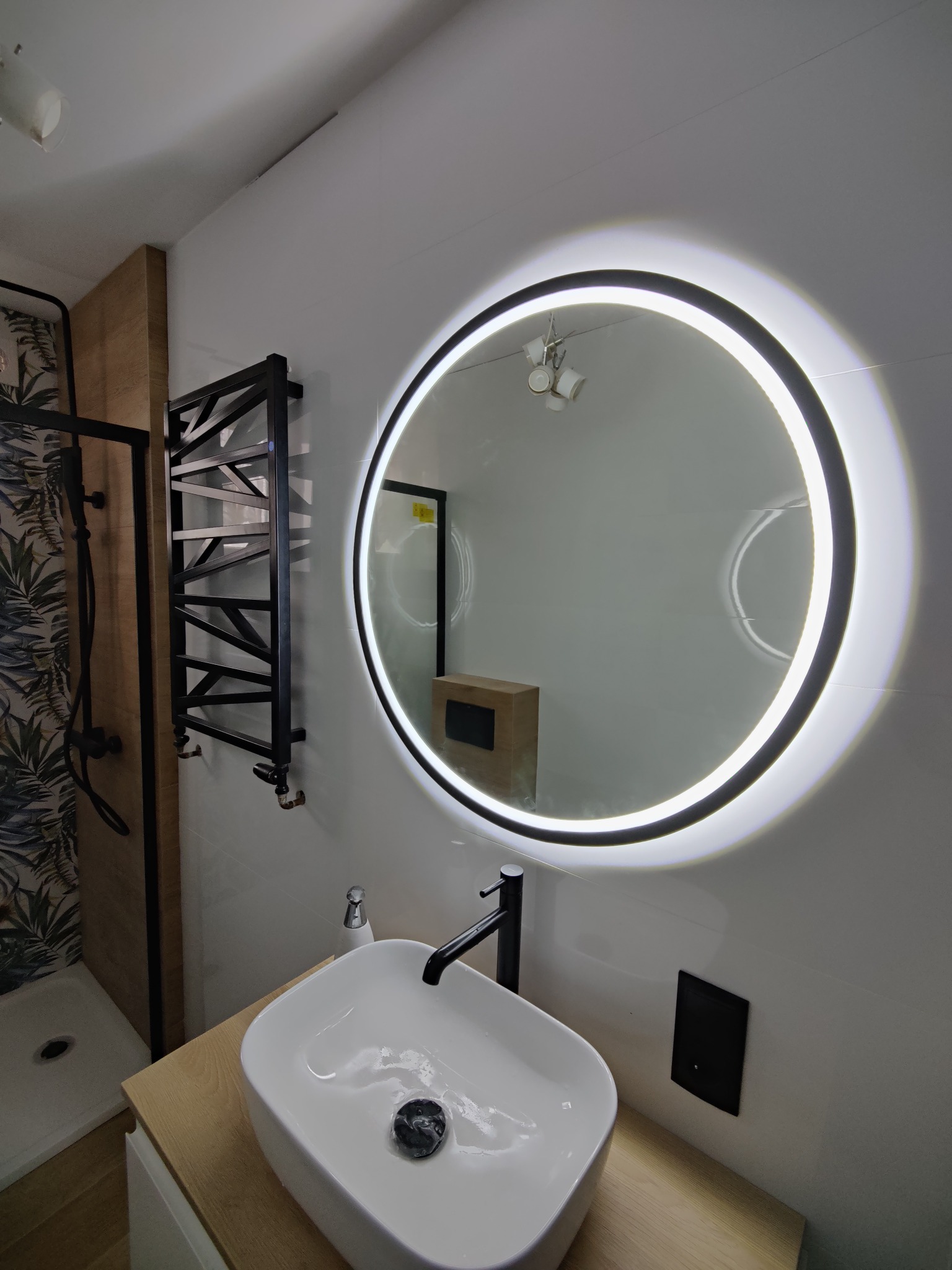 Nowoczesna łazienka z okrągłym podświetlanym lustrem, białą umywalką nablatową z czarną armaturą, czarnym grzejnikiem drabinkowym i kabiną prysznicową z motywem liści.
