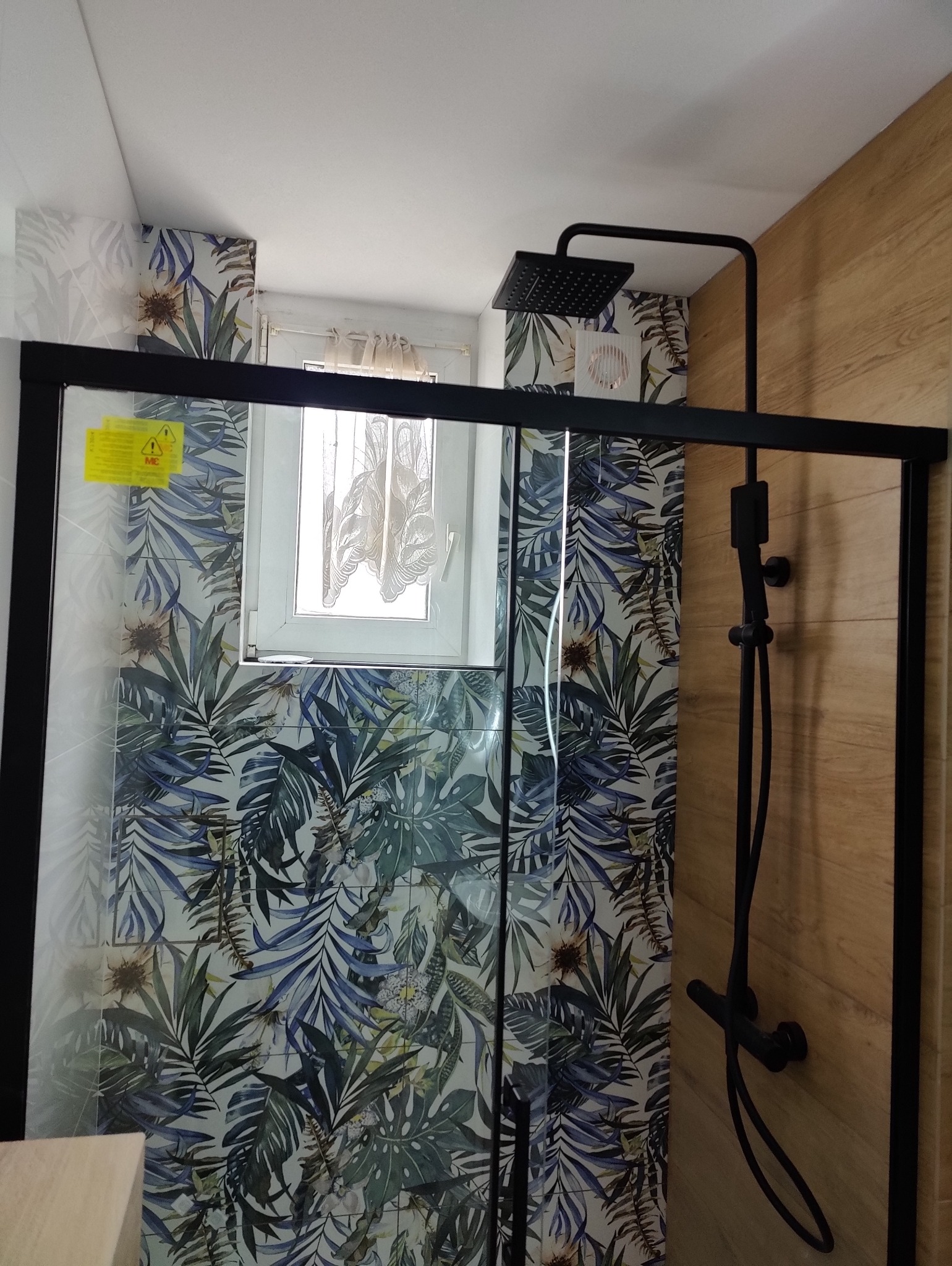 Nowoczesna kabina prysznicowa z czarną armaturą i deszczownicą, wykończona płytkami w motywy tropikalne i drewnopodobne, widoczne okno z zasłonką.
