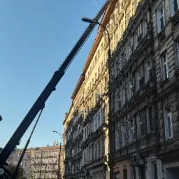 Wysięgnik podnośnika koszowego sięgający do elewacji kamienicy w trakcie prac, w tle widoczny ślad po przelatującym samolocie na błękitnym niebie.