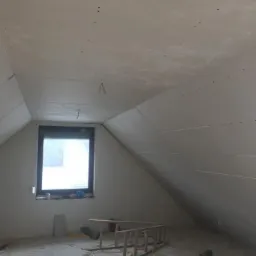 Poddasze ze skośnym sufitem pokryte płytami kartonowo-gipsowymi, widoczne ślady po szpachlowaniu, okno w czarnej ramie, drabina i narzędzia na podłodze.