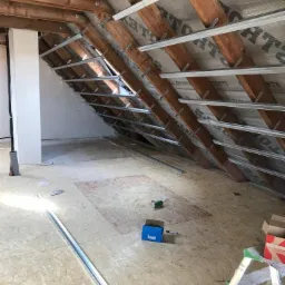 Poddasze w trakcie ocieplania, widoczne drewniane krokwie, folia paroizolacyjna, metalowe profile do montażu płyt kartonowo-gipsowych, częściowo ułożona podłoga z płyt OSB, pudełko Knauf i drabina.