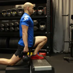 Trening funkcjonalny: mężczyzna wykonuje wykrok z kettlebell na stepie w siłowni, widoczne regały z obciążeniami i rower treningowy Assault Fitness.