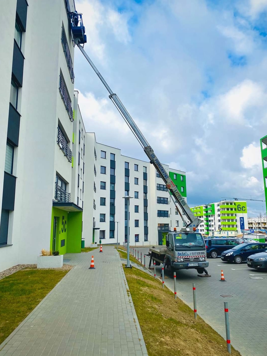 Podnośnik koszowy z pracownikiem przy białym budynku mieszkalnym z zielonymi akcentami, widoczne zaparkowane samochody i chodnik z pachołkami.