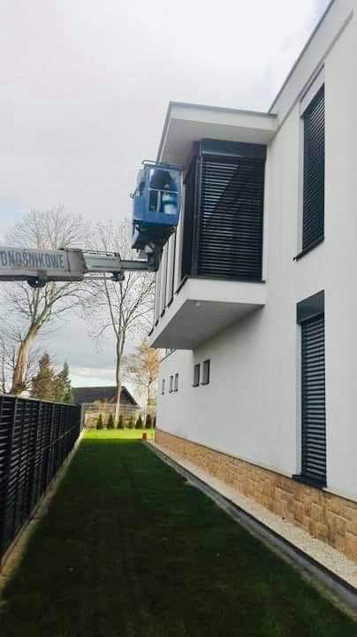 Podnośnik koszowy z pracownikiem przy oknie nowoczesnego domu z białymi ścianami i ciemnymi roletami zewnętrznymi, widok z poziomu trawnika.