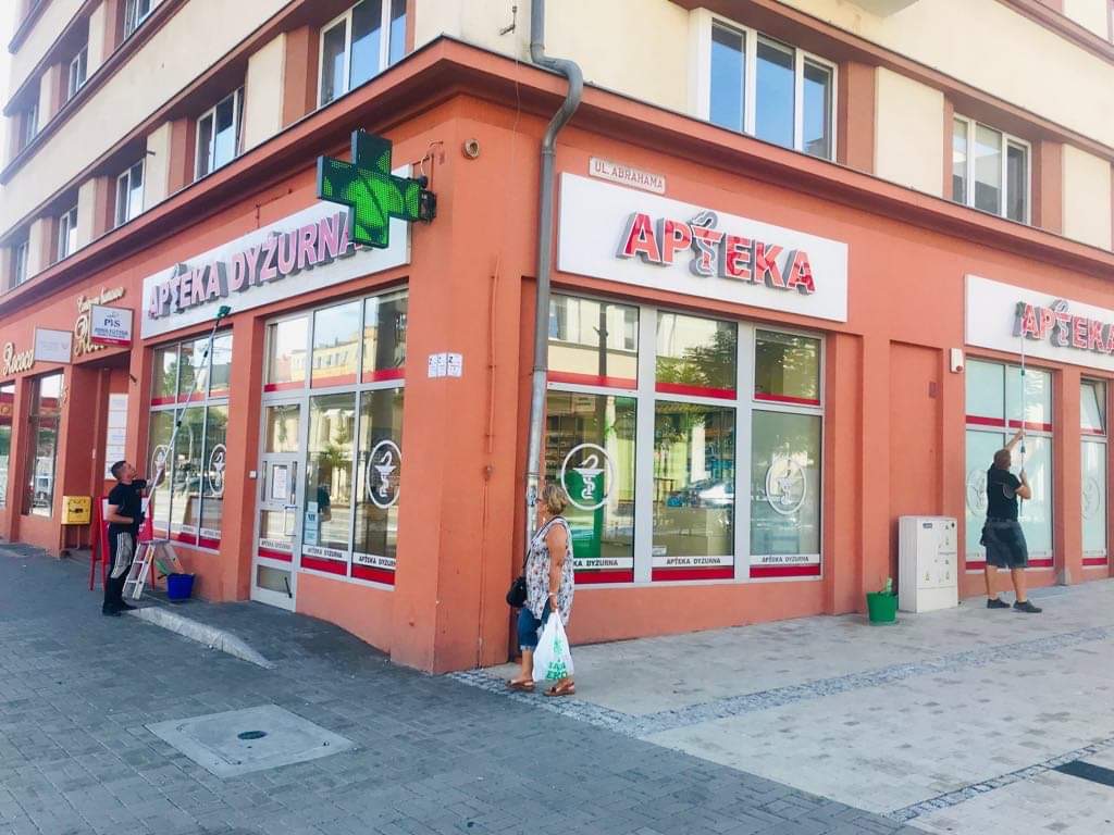 Mycie okien apteki przy ulicy Abrahama przez dwóch mężczyzn używających teleskopowych kijów i drabiny, widoczna kobieta z torbą na zakupy.