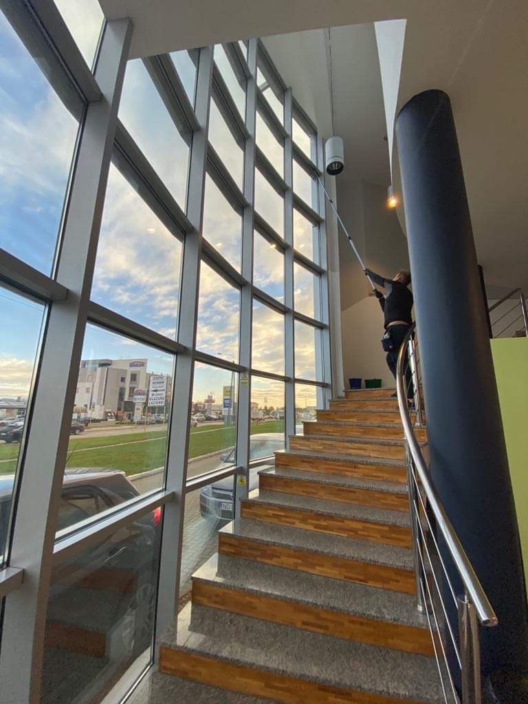 Pracownik na schodach czyści wysokie okna w budynku biurowym za pomocą teleskopowego kija z nakładką, widok na zewnątrz przez okna ukazuje budynki i parking.