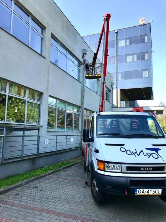 Pracownik na podnośniku koszowym czyści okna budynku biurowego, widoczny fragment elewacji, samochód dostawczy z podnośnikiem oraz budynek w tle.