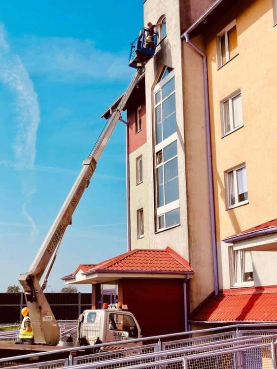 Pracownik w podnośniku koszowym czyści okno na wysokości w budynku wielorodzinnym, widoczny samochód dostawczy i pracownik w kamizelce odblaskowej na ziemi.