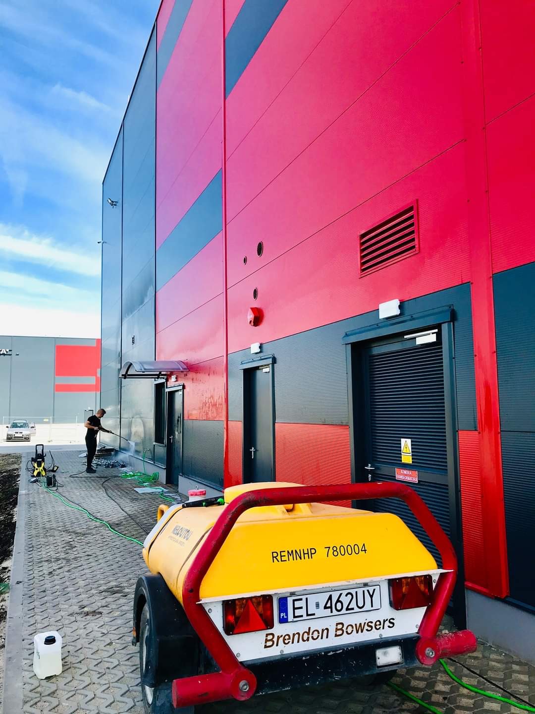 Czyszczenie elewacji przemysłowej z użyciem myjki ciśnieniowej; widoczny pracownik, fragment budynku w kolorach czerwonym i szarym oraz przyczepa z urządzeniem czyszczącym.